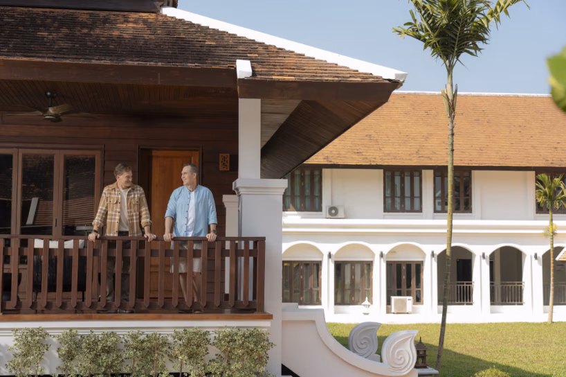 Two men talking outside a serene, traditional-style building surrounded by lush greenery and garden paths.